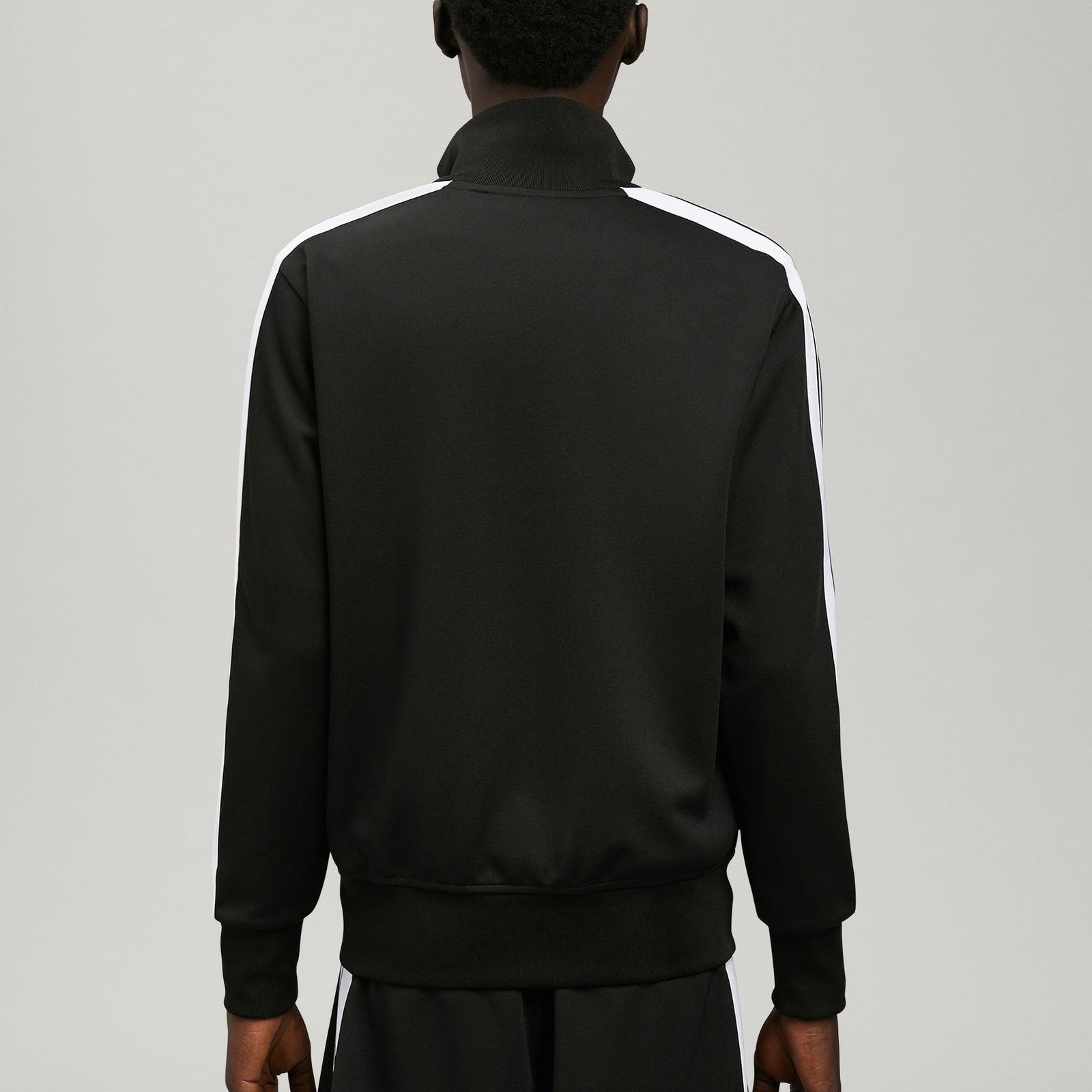 A man viewed from the back wearing a black track jacket with white shoulder stripes and black pants, photographed against a light gray studio background.