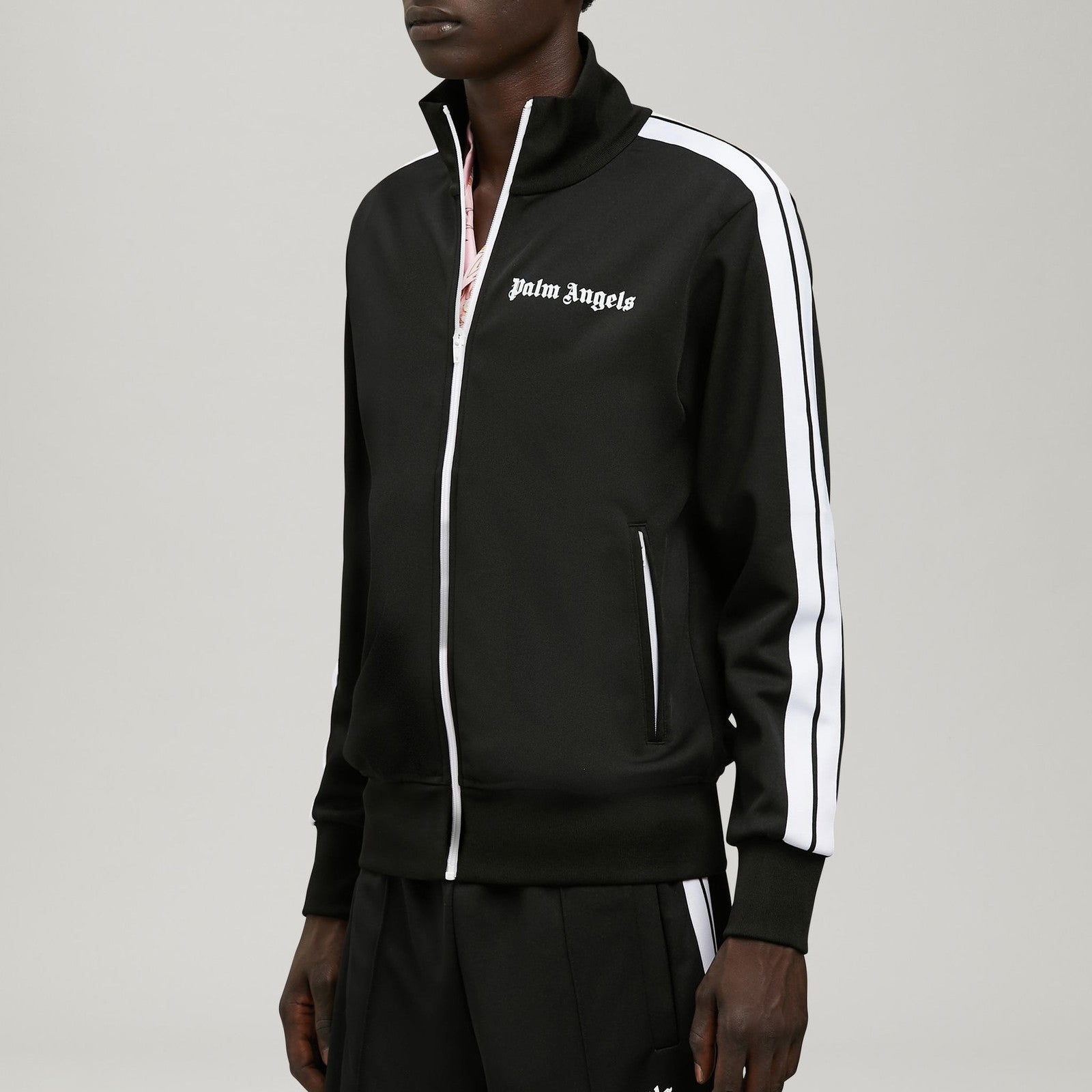 A model wearing a black Palm Angels track jacket with white zippers and white sleeve stripes, paired with matching black track pants, shot against a neutral gray background.