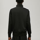 A man viewed from the back wearing a black track jacket with white shoulder stripes and black pants, photographed against a light gray studio background.