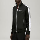 A model wearing a black Palm Angels track jacket with white zippers and white sleeve stripes, paired with matching black track pants, shot against a neutral gray background.